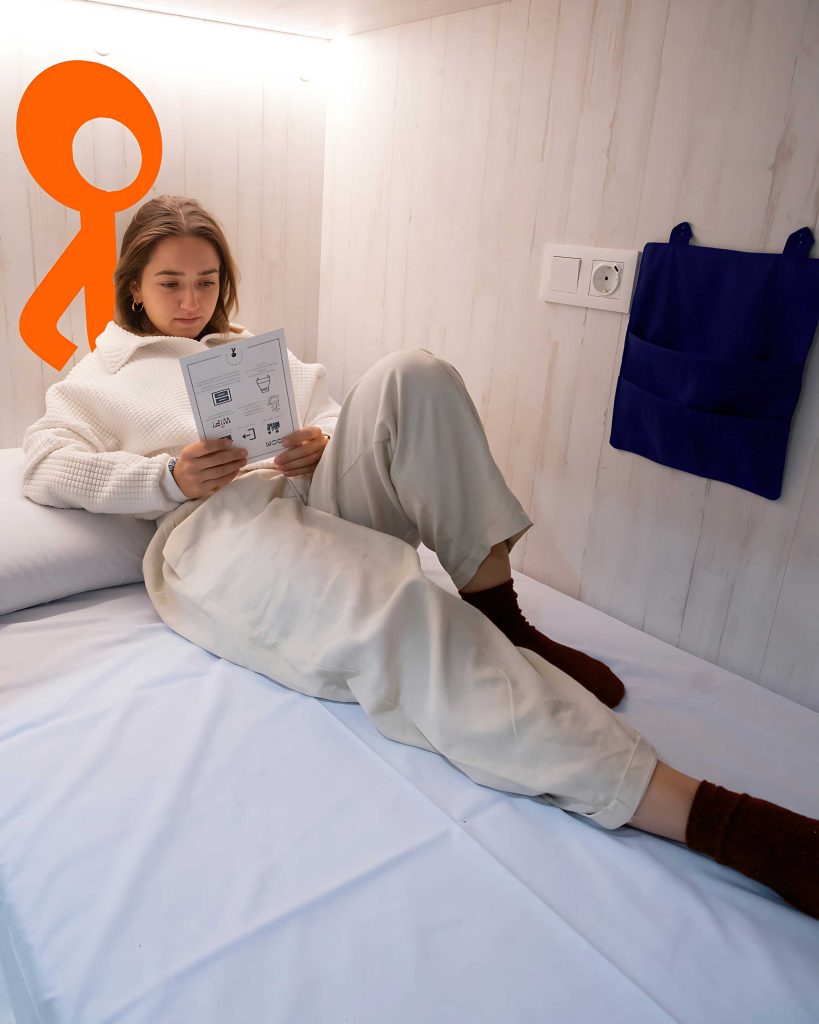 Mujer joven leyendo en la cama de una habitación minimalista de Adventure Hostel Madrid, con luz cálida y logo naranja en la pared.